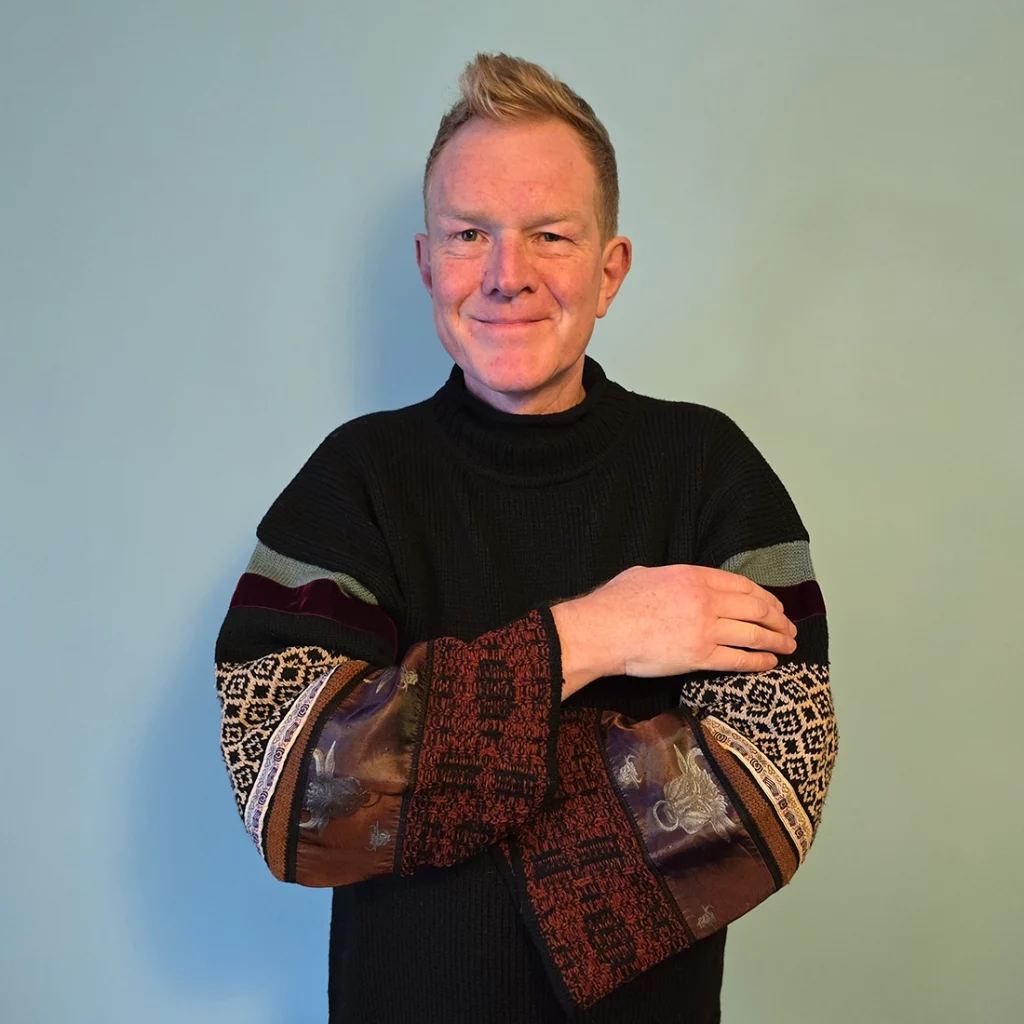 Person standing with arms crossed wearing multicoloured jumper