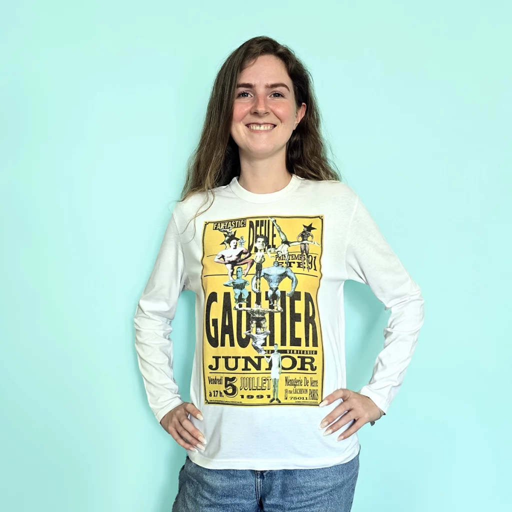 Model wearing a white Jean Paul Gaultier Junior long-sleeve shirt with a large yellow graphic print inspired by the Spring/Summer 1991 collection, featuring circus-style acrobatic figures, typography and oversized Gaultier Junior lettering.