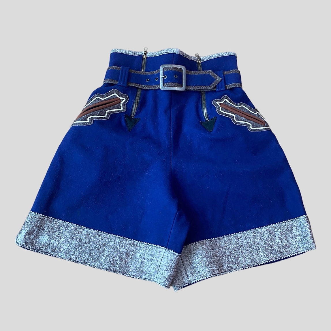 Royal blue lederhosen-style shorts with a wide belt, metal buckle, decorative curved pockets with contrasting trim, and pale blue patterned fabric along the hem.